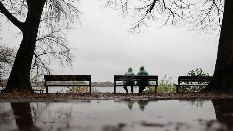 Zwei Menschen mit grünen Regenponchos sitzen bei leichtem Dauerregen auf einer Bank an der Außenalster am 14.11.2025 in Hamburg.