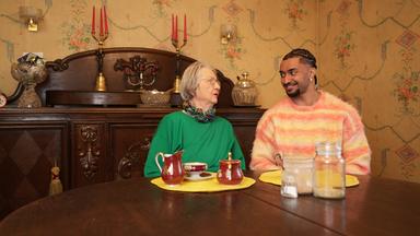 Eva (81) und Louis (26) sitzen an einem Tisch und schauen sich vertraut an. Im Hintergrund steht eine antike Anrichte.