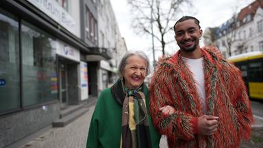 Oma Eva und Enkel Louis spazieren Arm in Arm eine Straße entlang und lächeln in die Kamera.