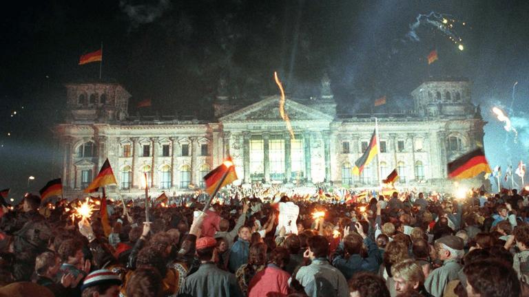 Am 3. Oktober 1990 feiern jubelnde Menschen vor dem Berliner Reichstag mit Feuerwerk, Deutschlandfahnen und Volksfeststimmung die wiedergewonnene Einheit Deutschlands.