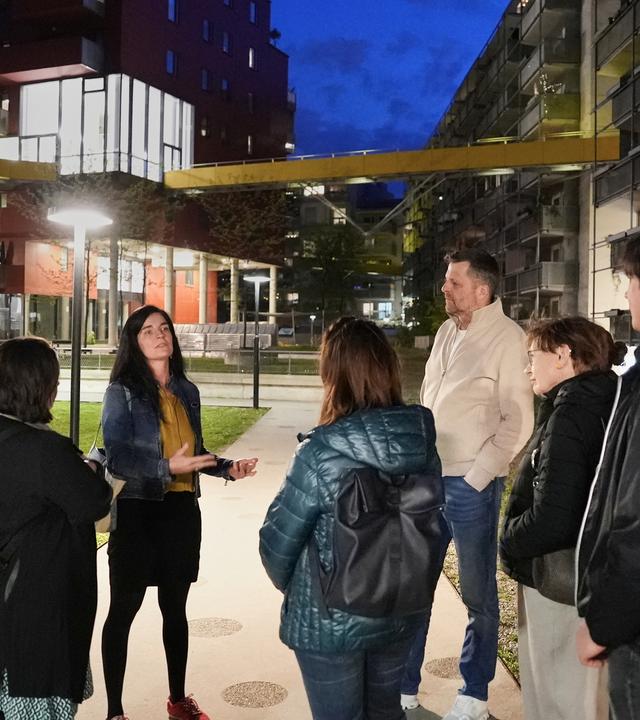Julia Girardi-Hoog steht mit fünf Personen bei einer nächtlichen Führung im Wiener Sonnwendviertel. Sie stehen an einem Grünstreifen zwischen mehrstöckigen Gebäuden, der mit Straßenlaternen hell beleuchtet ist.