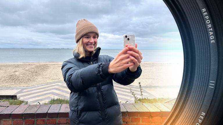 Ariana Müller steht auf der Strandpromenade, hinter ihr liegt der leere Strand und das Meer. Sie nimmt gerade ein Video mit dem Smartphone auf.