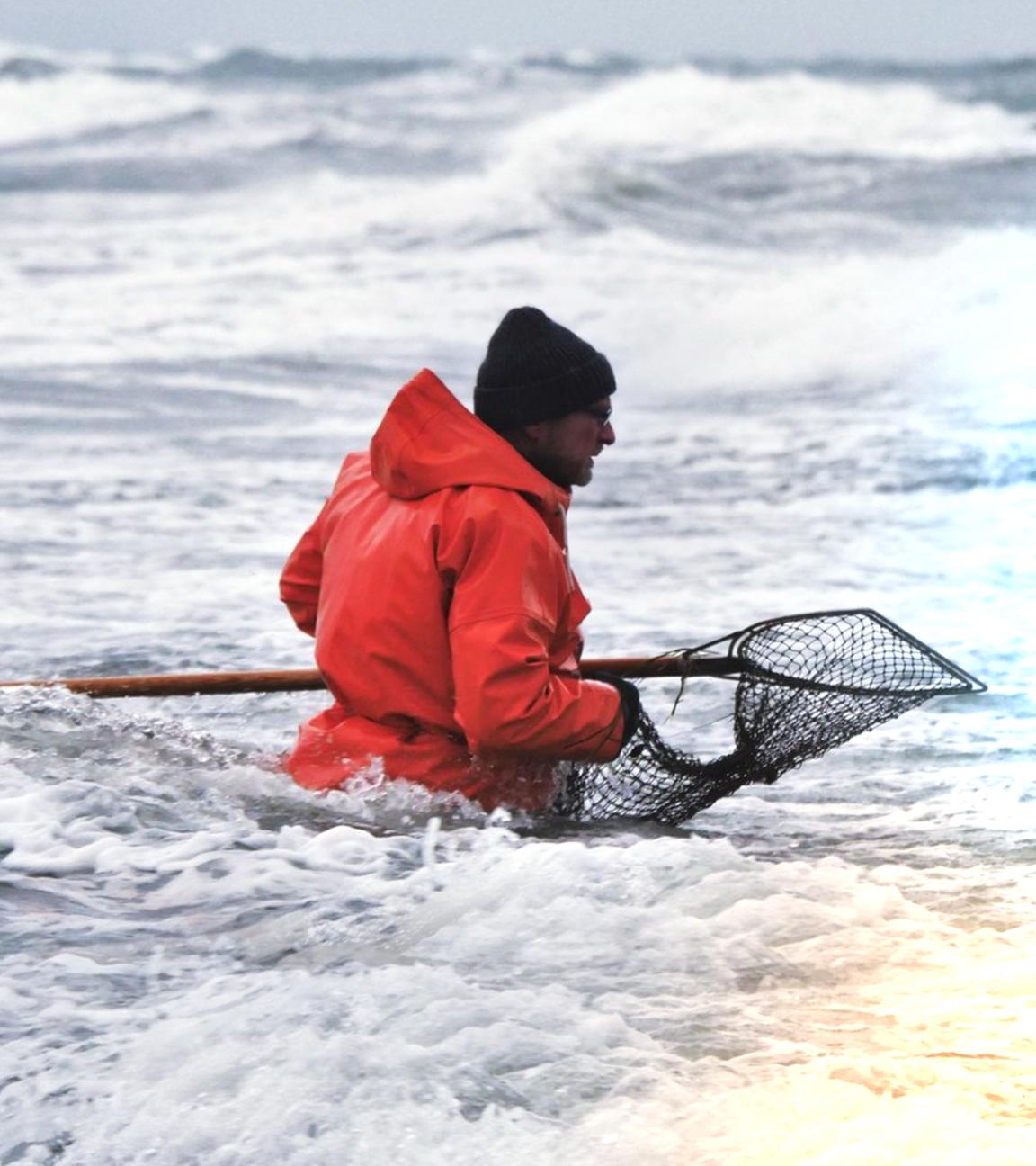Ein Mann in knallroter Jacke hält einen großen Kescher und steht bis zur Hüfte im unruhigen Meer.
