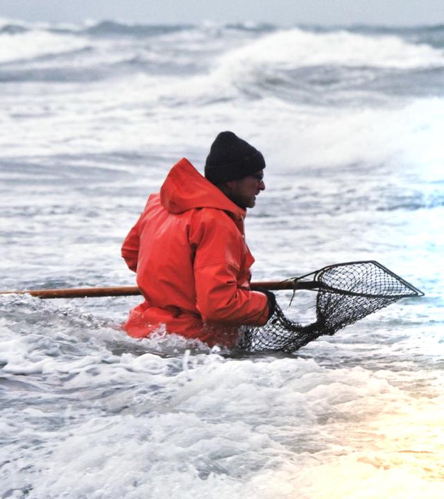 Ein Mann in knallroter Jacke hält einen großen Kescher und steht bis zur Hüfte im unruhigen Meer.