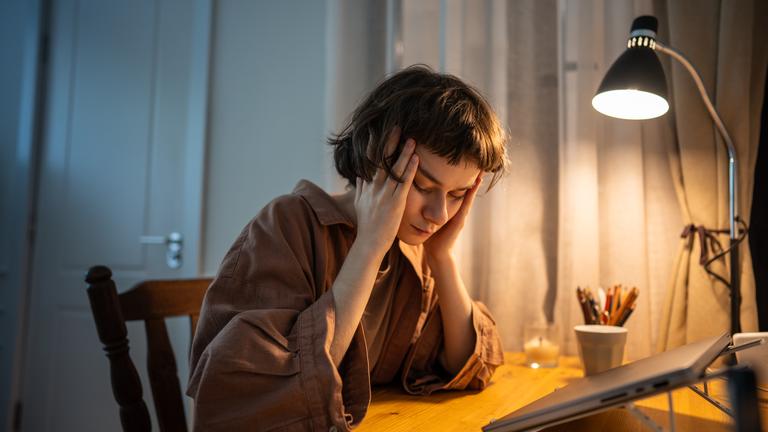 Eine müde junge Frau reibt sich die Schläfen. Sie sitzt am Schreibtisch mit dem Kopf in ihren Händen. Ihre Schreibtischlampe brennt.