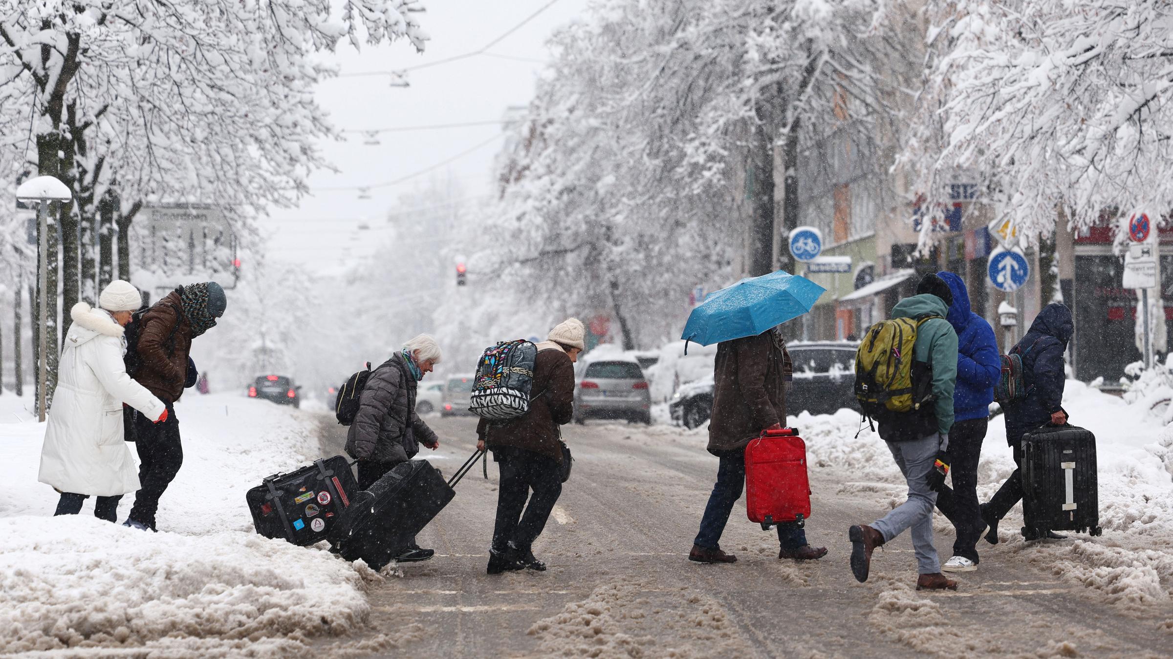 Eine Gruppe von Menschen mit ihrem Gepäck überquert eine schneebedeckte Straße in der Nähe des Münchner Hauptbahnhofs in München, Deutschland, 02.12.2023.