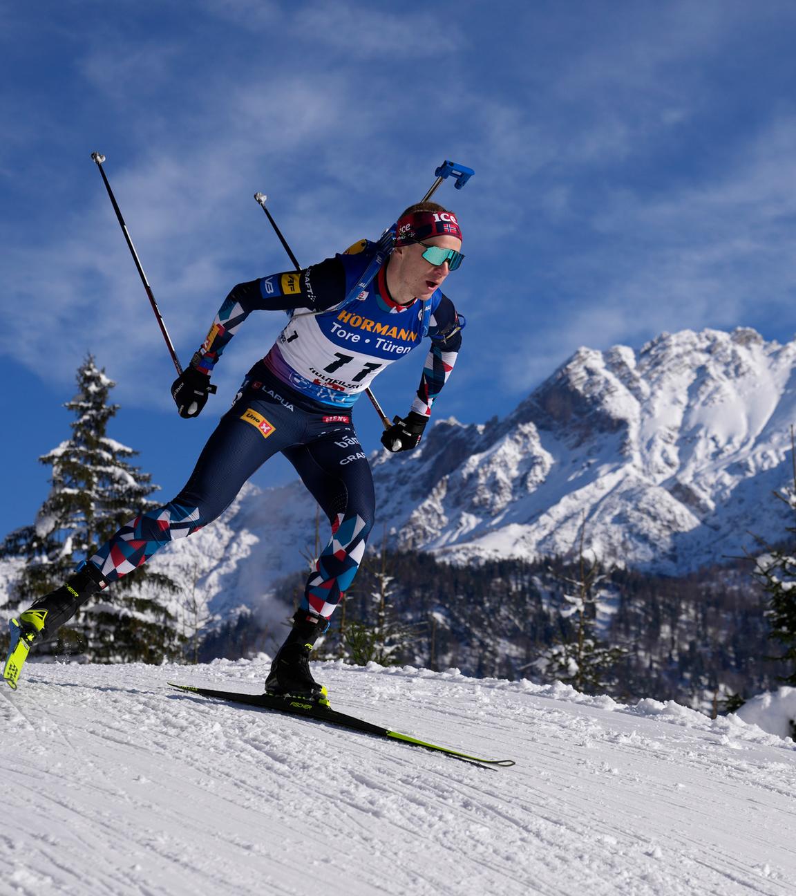 Ein Biathlon-Sportler auf einer Piste mit verschneiten Bergen im Hintergrund.