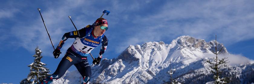 Ein Biathlon-Sportler auf einer Piste mit verschneiten Bergen im Hintergrund.