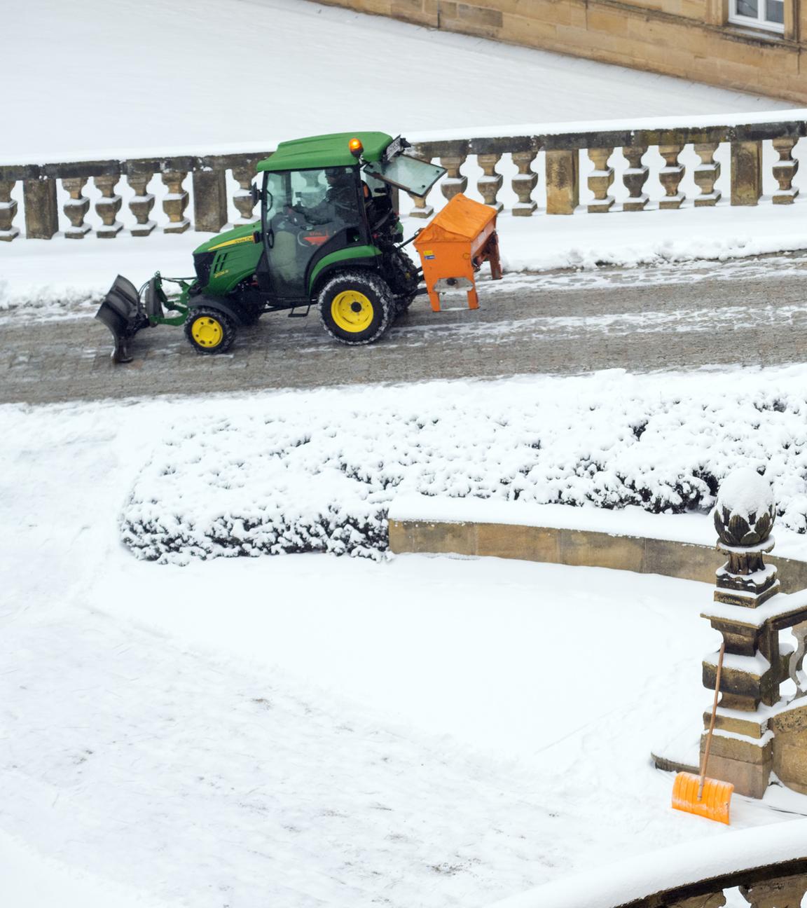 12.01.2026, Bayern, Neubanz: Eine Schneeschaufel lehnt an einem historischen Treppengeländer, während ein Räumfahrzeug Salz auf einem Weg im Innenhof von Kloster Banz streut. 