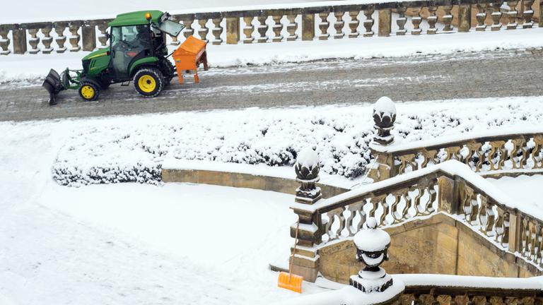 12.01.2026, Bayern, Neubanz: Eine Schneeschaufel lehnt an einem historischen Treppengeländer, während ein Räumfahrzeug Salz auf einem Weg im Innenhof von Kloster Banz streut. 