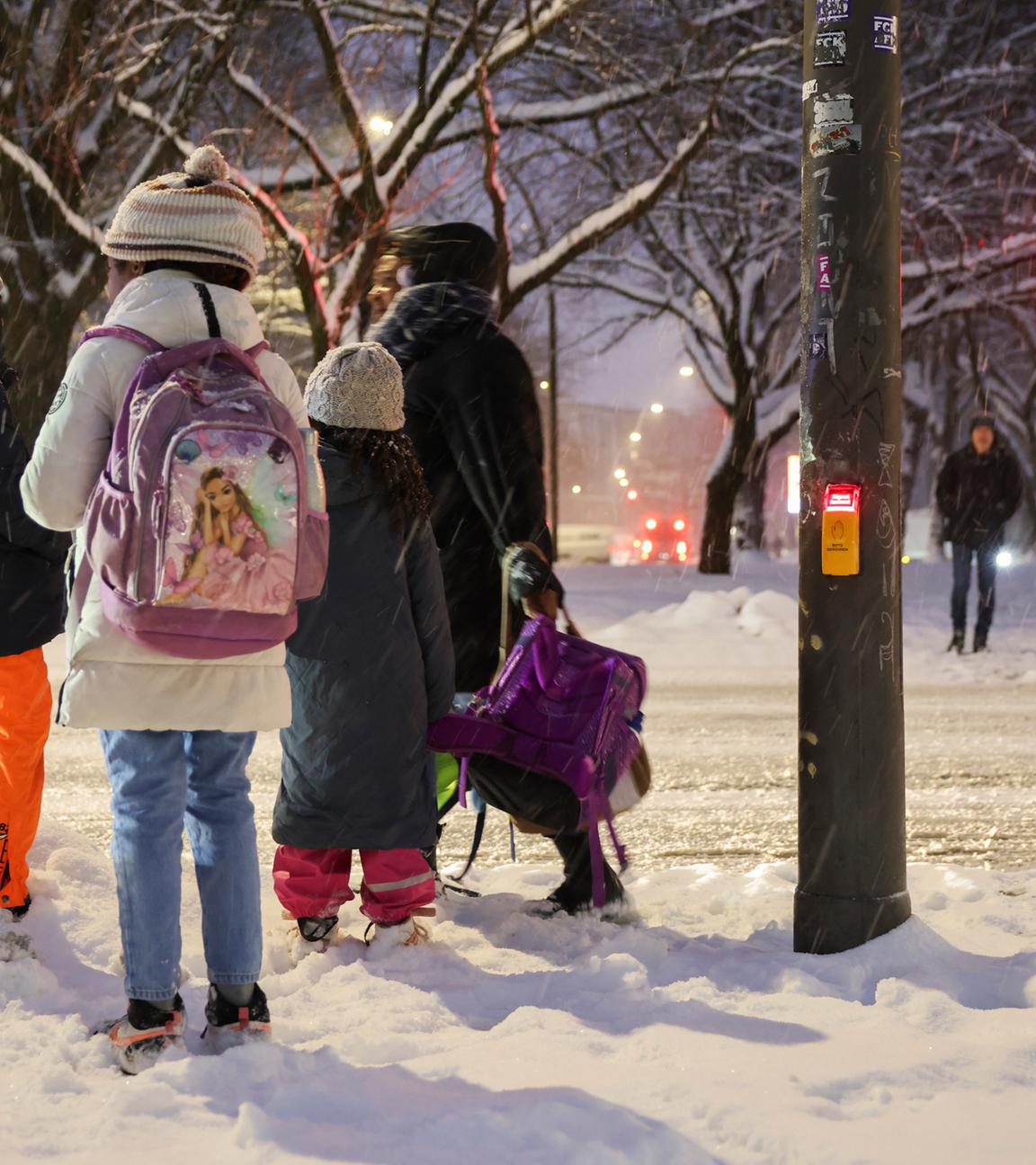 Eine Mutter trägt die Schulranzen ihrer Kinder während sie im Schnee an einer Fußgängerampel über die Kieler Straße warten.