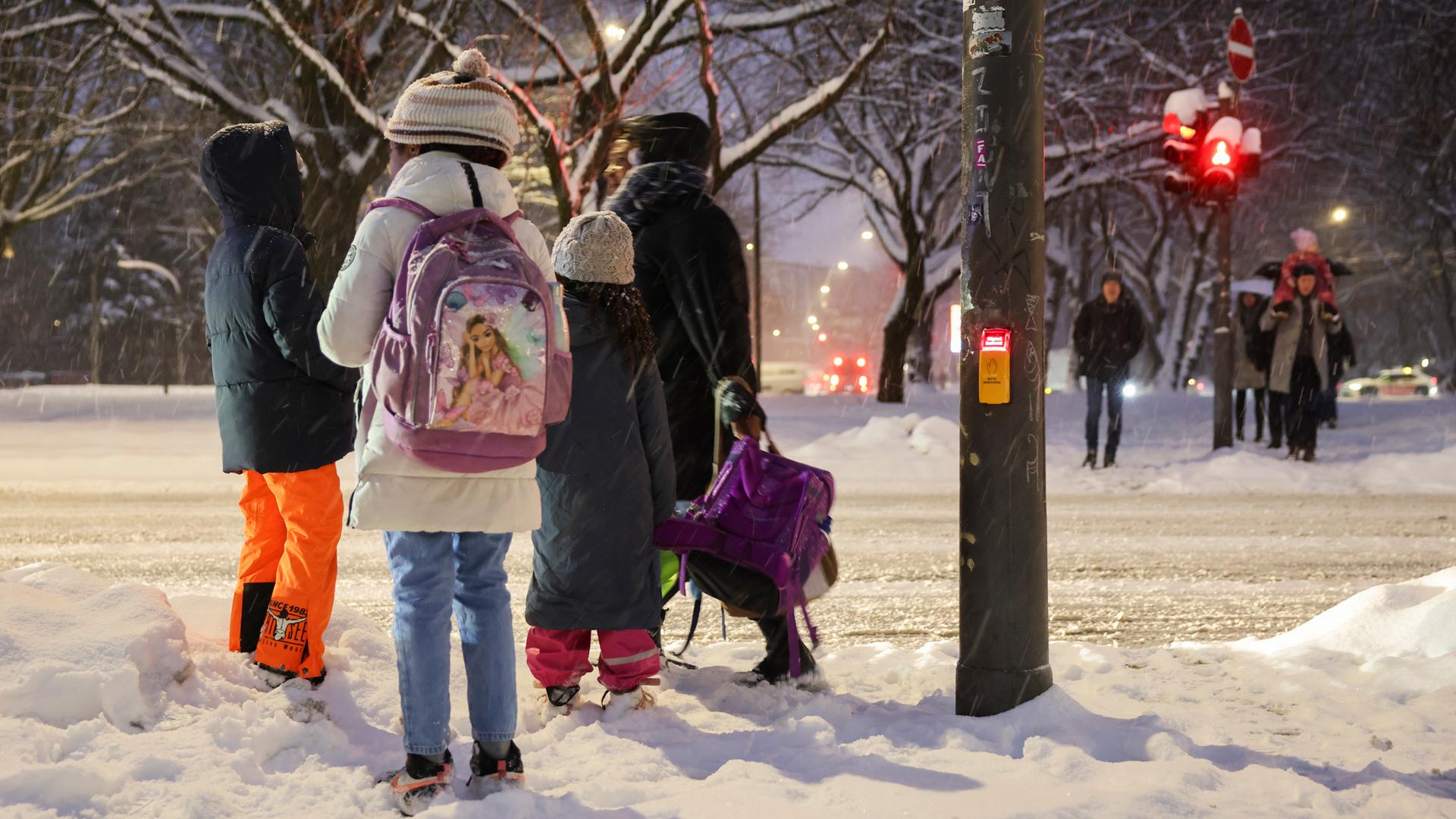 Eine Mutter trägt die Schulranzen ihrer Kinder während sie im Schnee an einer Fußgängerampel über die Kieler Straße warten.
