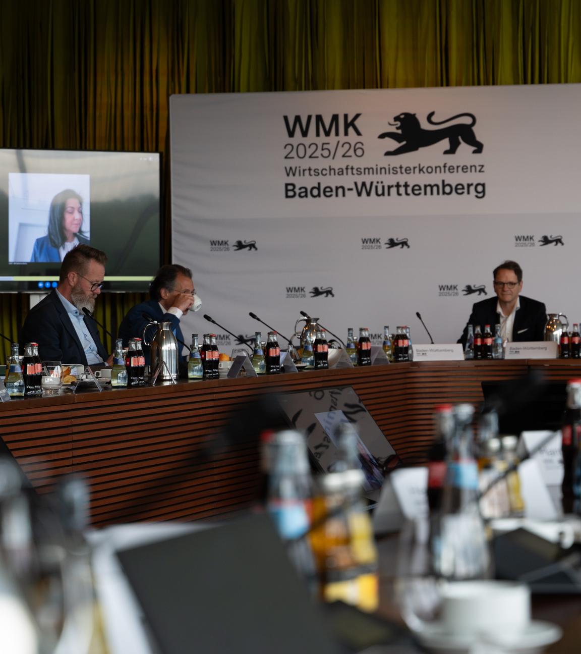 Konferenzraum mit Teilnehmern hinten an den Tischen vor Mikrofonen, Katherina Reiche ist über einen Bildschirm zugeschaltet.