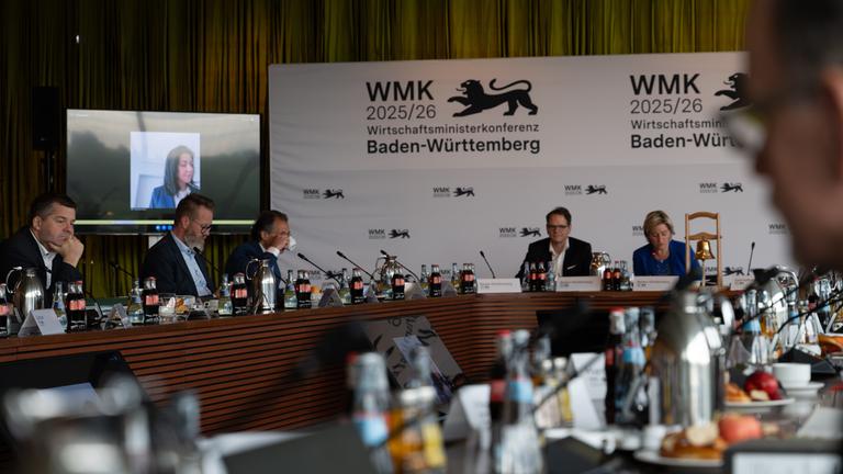 Konferenzraum mit Teilnehmern hinten an den Tischen vor Mikrofonen, Katherina Reiche ist über einen Bildschirm zugeschaltet.