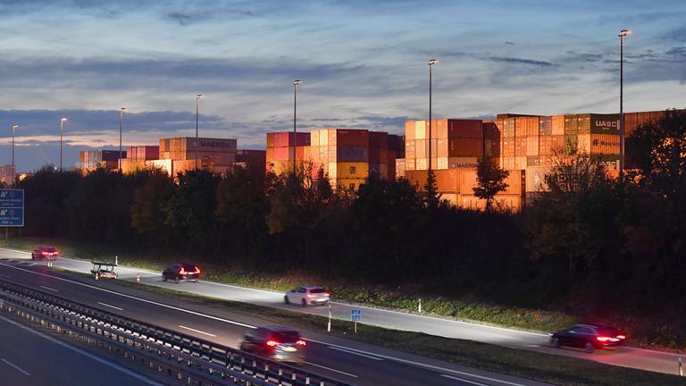 Autobahn bei Sonnenuntergang, dahinter stehen gestapelte Container.