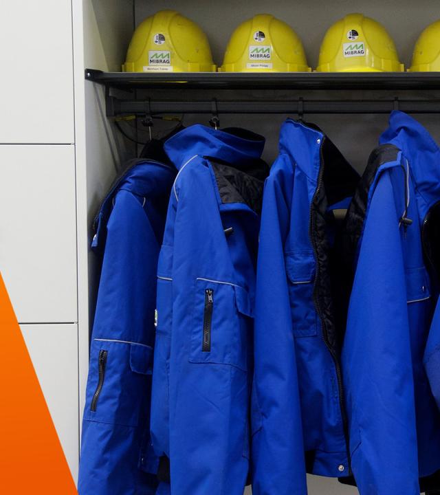 Ein Schrank mit blauen Arbeitsanzügen und oben drüber ein Fach mit Helmen
