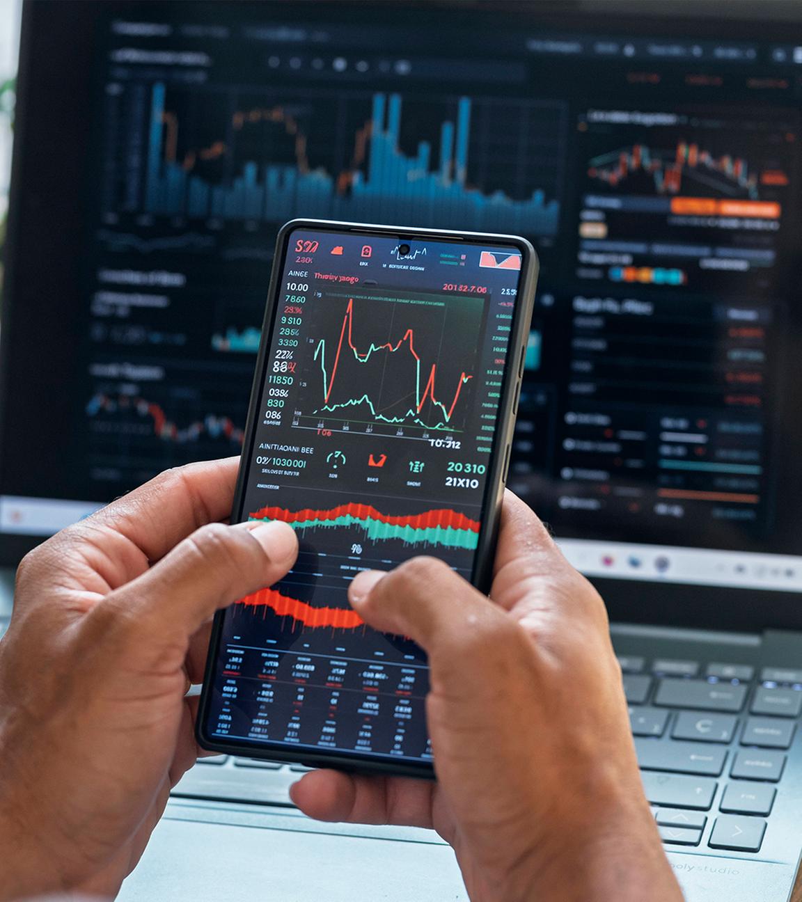 Notebbook-Bildschirm mit Börsenkursen. Dabor hält ein Mann ein Handy mit Börsenkursen.