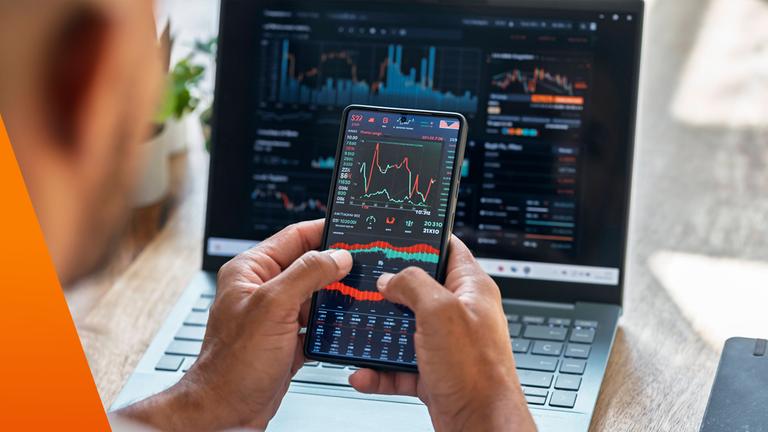 Notebbook-Bildschirm mit Börsenkursen. Dabor hält ein Mann ein Handy mit Börsenkursen.