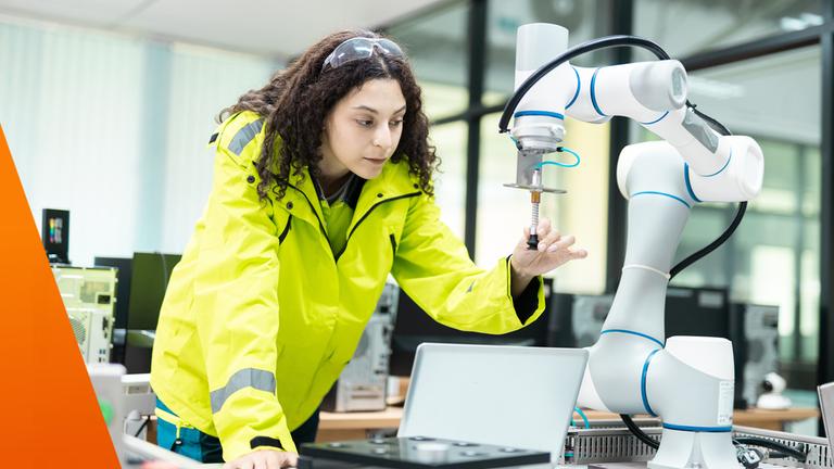 Eine Frau in einer Werkhalle oder einem Labor beugt sich über einen Tisch und arbeitet konzentriert an einem weißen Robotarm mit Greifwerkzeug. Sie trägt eine auffällige gelbe Schutzjacke und blickt auf ein Bauteil, das der Roboter hält. Neben ihr steht ein Laptop, im Hintergrund sind technische Geräte zu sehen. Das Bild zeigt die Entwicklung und Anwendung moderner Robotik.