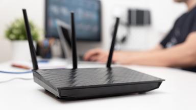 Ein Wlan-Router steht auf einem Tisch vor einer am PC arbeitenden Person.