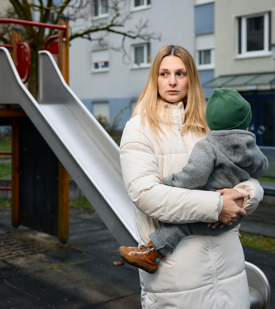Annie hält ihr Baby auf dem Arm und schaut traurig und erschöpft zur Seite. Im Hintergrund ist ein Spielplatz mit einer Rutsche zu sehen.