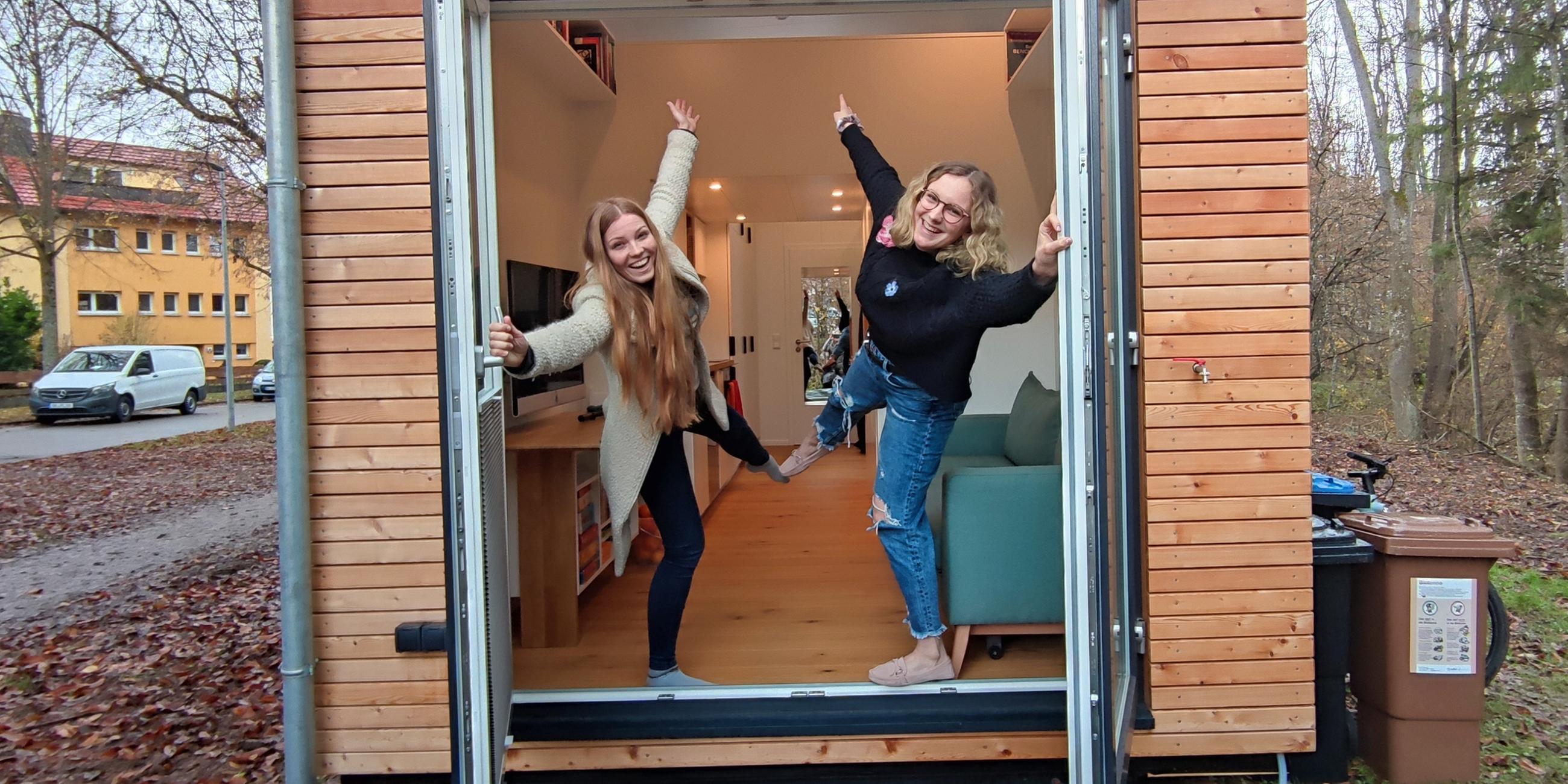 Reporterin Wiebke Denker und Joey stehen an der Tür eines Tiny-House. Sie strecken jede ein Bein und einen Arm in die Luft. Mit den anderen Armen halten sie sich an den geöffneten Türen fest. Beide lachen.
