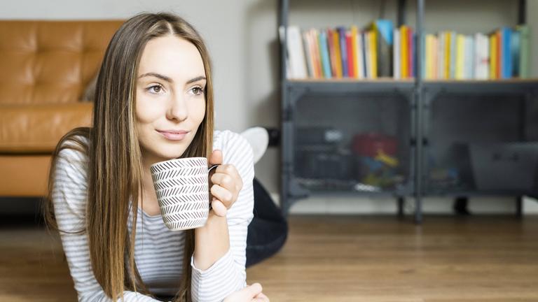 Entspannte junge Frau, die zu Hause auf dem Boden liegt und Kaffee trinkt.