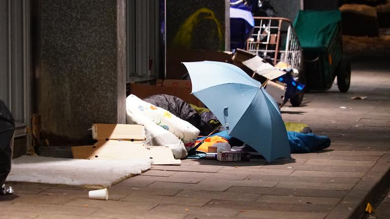 Ein Lager aus Matratzen und einem Regenschirm auf dem Gehweg neben einer Hauswand.