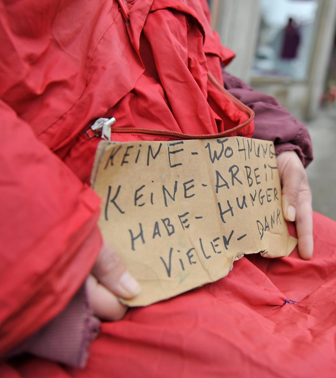Ein Bettler mit einem Schild ist zu sehen. Es wird um Geld gebeten und gesagt, dass die Person wohnungslos ist.