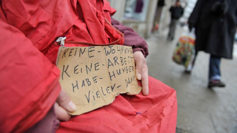 Ein Bettler mit einem Schild ist zu sehen. Es wird um Geld gebeten und gesagt, dass die Person wohnungslos ist.