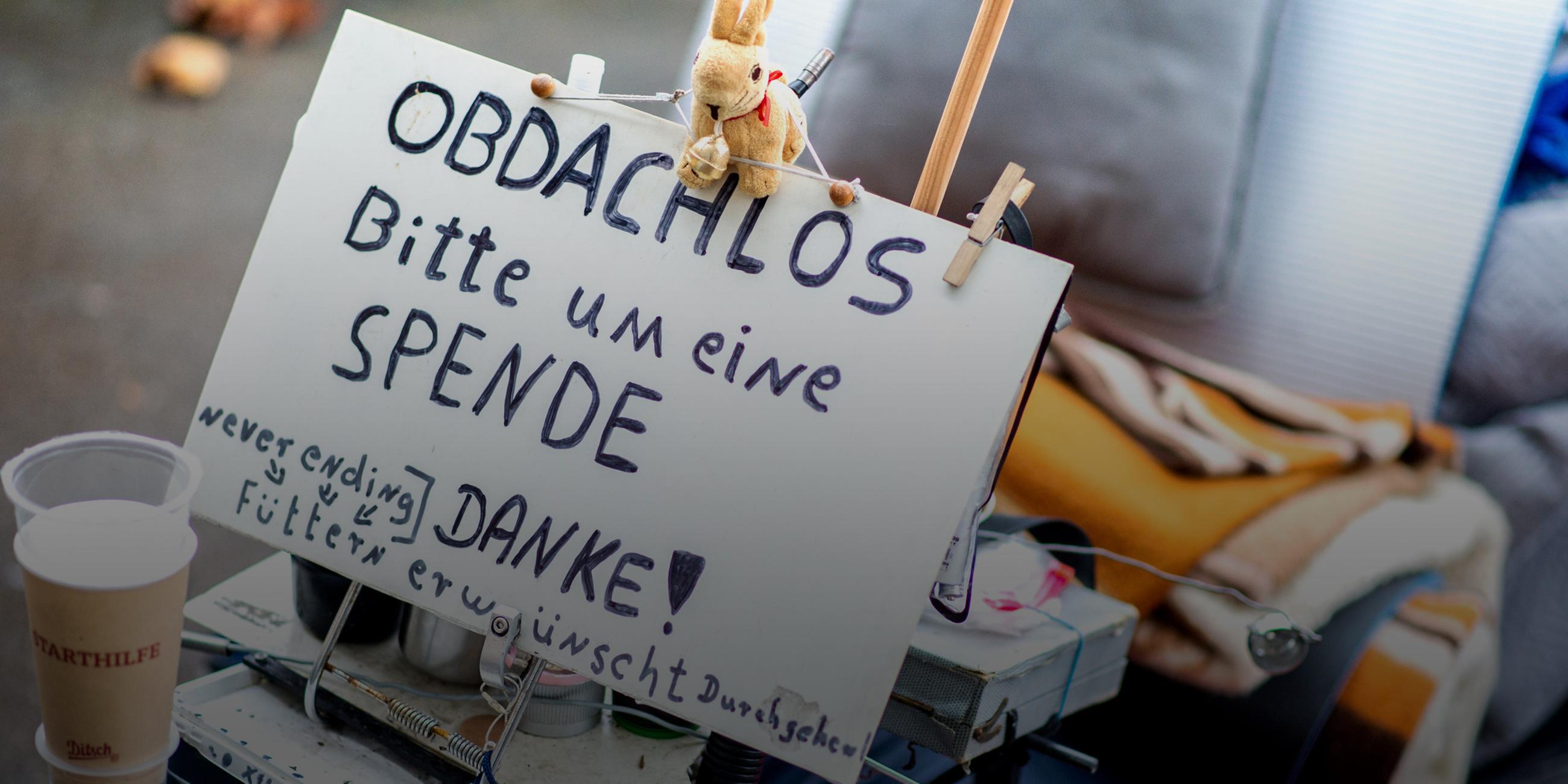 Ein Schild mit der Aufschrift ·Obdachlos - bitte um eine Spende - danke!· steht neben den Habseligkeiten eines Obdachlosen, die auf einem Bürgersteig in der Innenstadt liegen