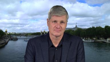 Spanien-Korrespondent Thomas Walde. Er trägt ein schwarzes Hemd und ein dunkelblaues Sakko. Er befindet sich in einem Studio. Der Hintergrund ist eine Aufnahme von einem Fluss in einer Stadt.