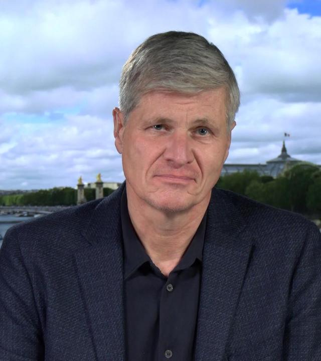 Spanien-Korrespondent Thomas Walde. Er trägt ein schwarzes Hemd und ein dunkelblaues Sakko. Er befindet sich in einem Studio. Der Hintergrund ist eine Aufnahme von einem Fluss in einer Stadt.