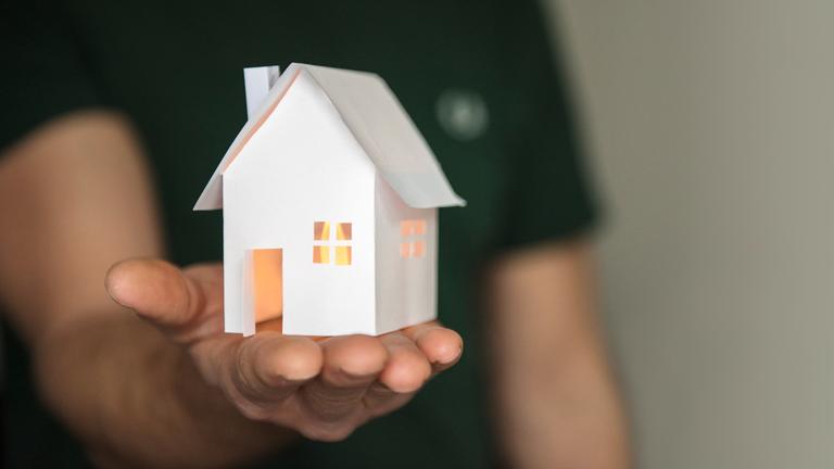Symbolhafter Wohungstausch: Ein Mann hält ein Einfamilienaus aus Papier in der Richtung Kamera ausgestreckten Hand.