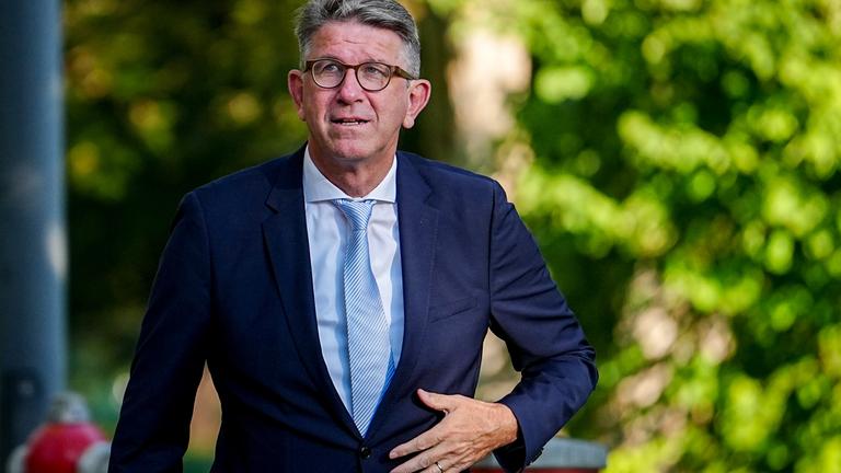 Ein Mann mit graumelierten kurzen Haaren, Brille und im dunkelblauen Anzug mit hellblauem Hemd und Krawatte fasst sich im Laufen mit der linken Hand vorne ans Jackett.