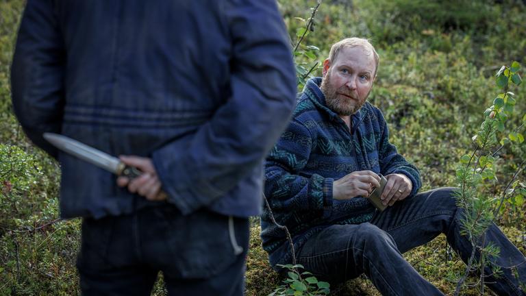 Ein Mann steht mit dem Rücken zur Kamera und hält ein Messer hinter dem Rücken, während vor ihm ein Mann sitzt und ihn anschaut.