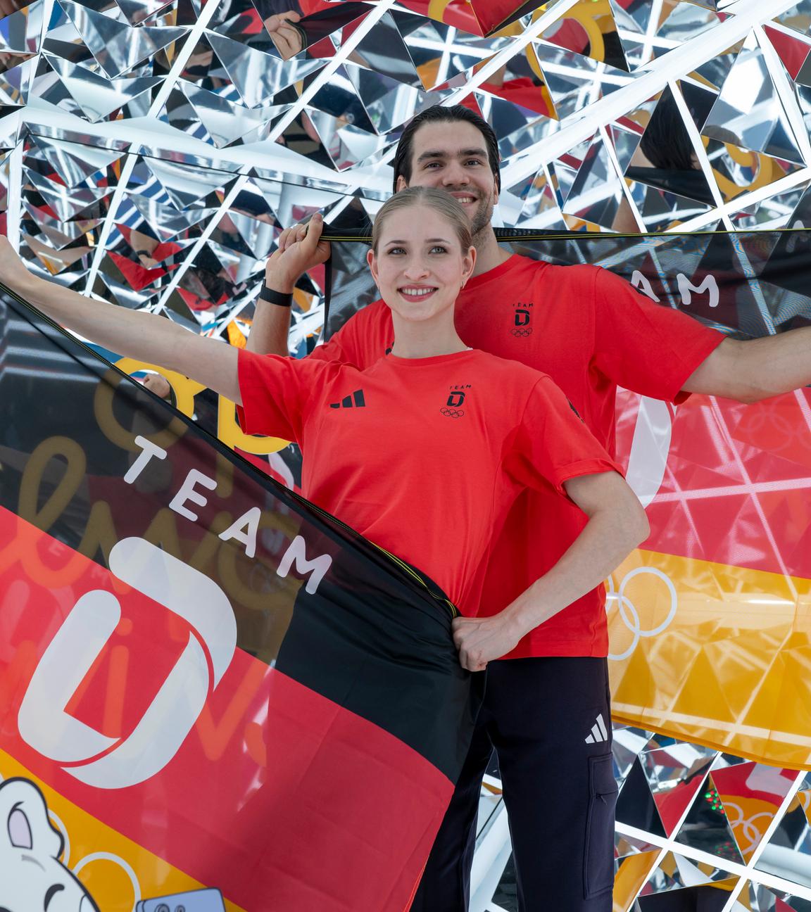 Die deutsche Eiskunstläuferin Minerva Fabienne Hase und der deutsche Eiskunstläufer Nikita Volodin präsentieren vor Glitzerkulisse die deutsche Flagge