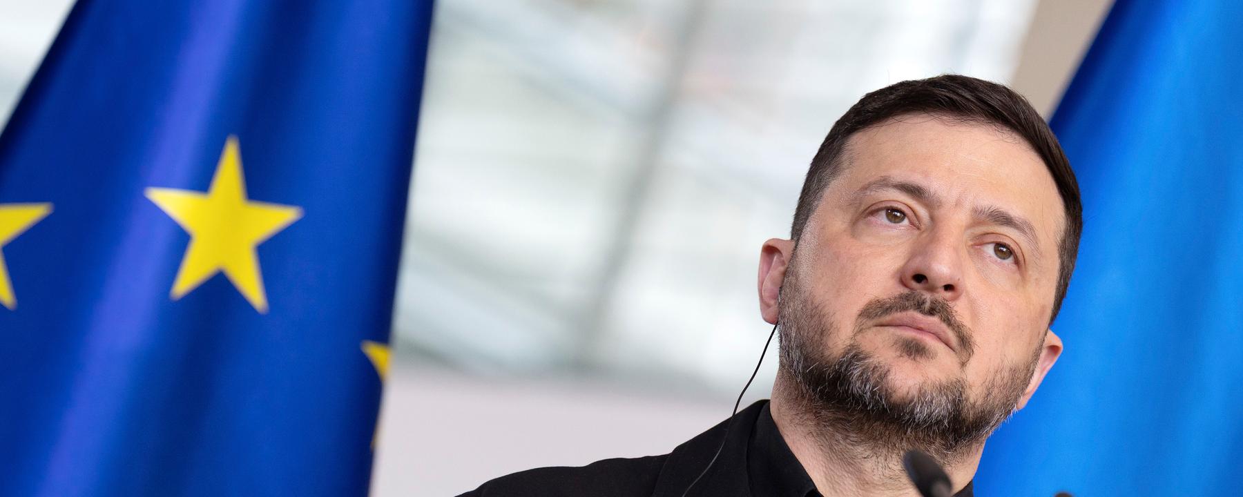 Wolodymyr Selenskyj steht vor einem Mikrofon-Pult. Im Hintergrund ist due Flagge der Europäischen Union zusehen.