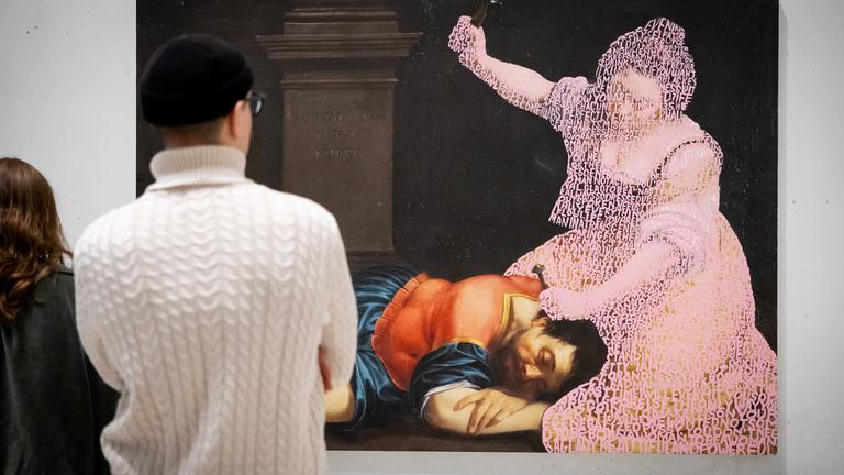 Ein Paar schaut sich ein großformatiges Bild an, auf dem ein Mann in historischer Kleidung schlafend auf dem Boden liegt, während eine Frau, die nur als verwitterter Umriss zu sehen ist, über ihm kniet und mit einer Hand ausholt.