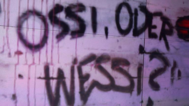 An einer Wand steht "OSSI ODER WESSI?", eine Studiodeko der ZDF-Talkshow "maybrit illner".
