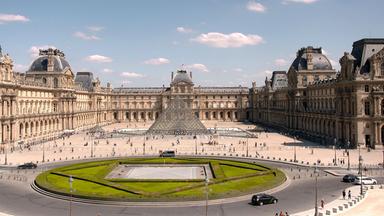 Ein Bauwerk umrandet einen Platz. In der Mitte des Platzes ragt eine gläserne Pyramide aus der Erde: Der Louvre in Paris. Um eine kreisförmige Grasfläche führt eine Straße.
