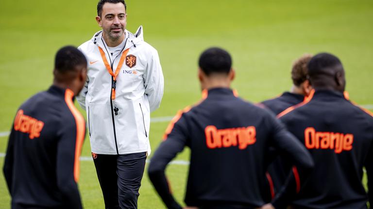 Niederländischer Trainer steht mit weißer Jacke vor Spielern die mit "Oranje" angeschrieben sind.
