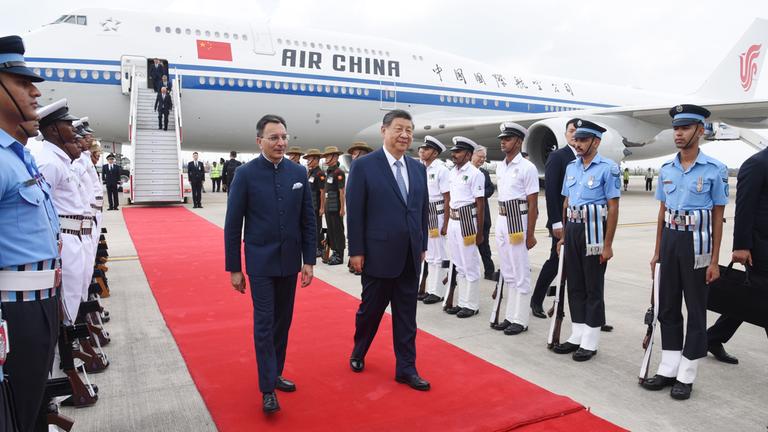 Der chinesische Präsident Xi Jinping läuft auf einem roten Teppich, im Hintergrund ist ein Flugzeug von Air China zu sehen.