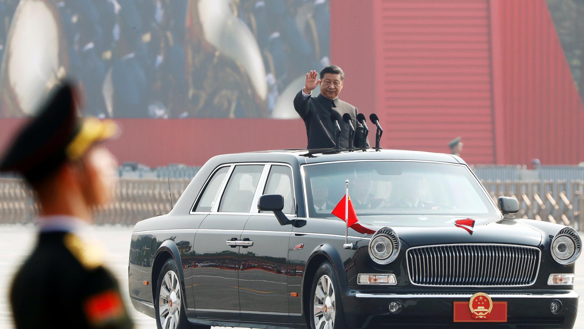Der chinesische Staatsminister Xi Jinping schaut aus dem Dach eines fahrenden Autos und winkt. Mit dem Auto fährt er durch eine Militärparade. Im Vordergrund ist verschwommen ein Mann in Uniform zu sehen.