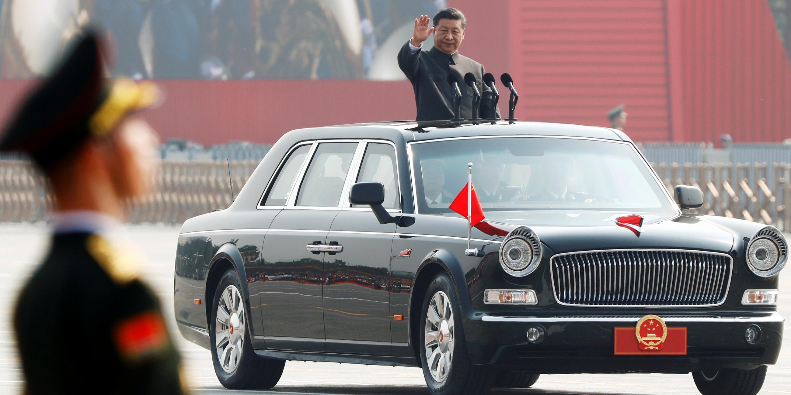 Der chinesische Staatsminister Xi Jinping schaut aus dem Dach eines fahrenden Autos und winkt. Mit dem Auto fährt er durch eine Militärparade. Im Vordergrund ist verschwommen ein Mann in Uniform zu sehen.