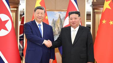 Xi Jinping und Kom Jong Un stehen vor Chinas und Nordkoreas Fahnen und schütteln die Hände.