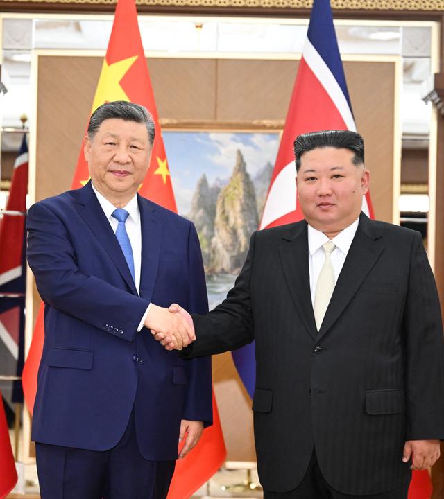 Xi Jinping und Kom Jong Un stehen vor Chinas und Nordkoreas Fahnen und schütteln die Hände.