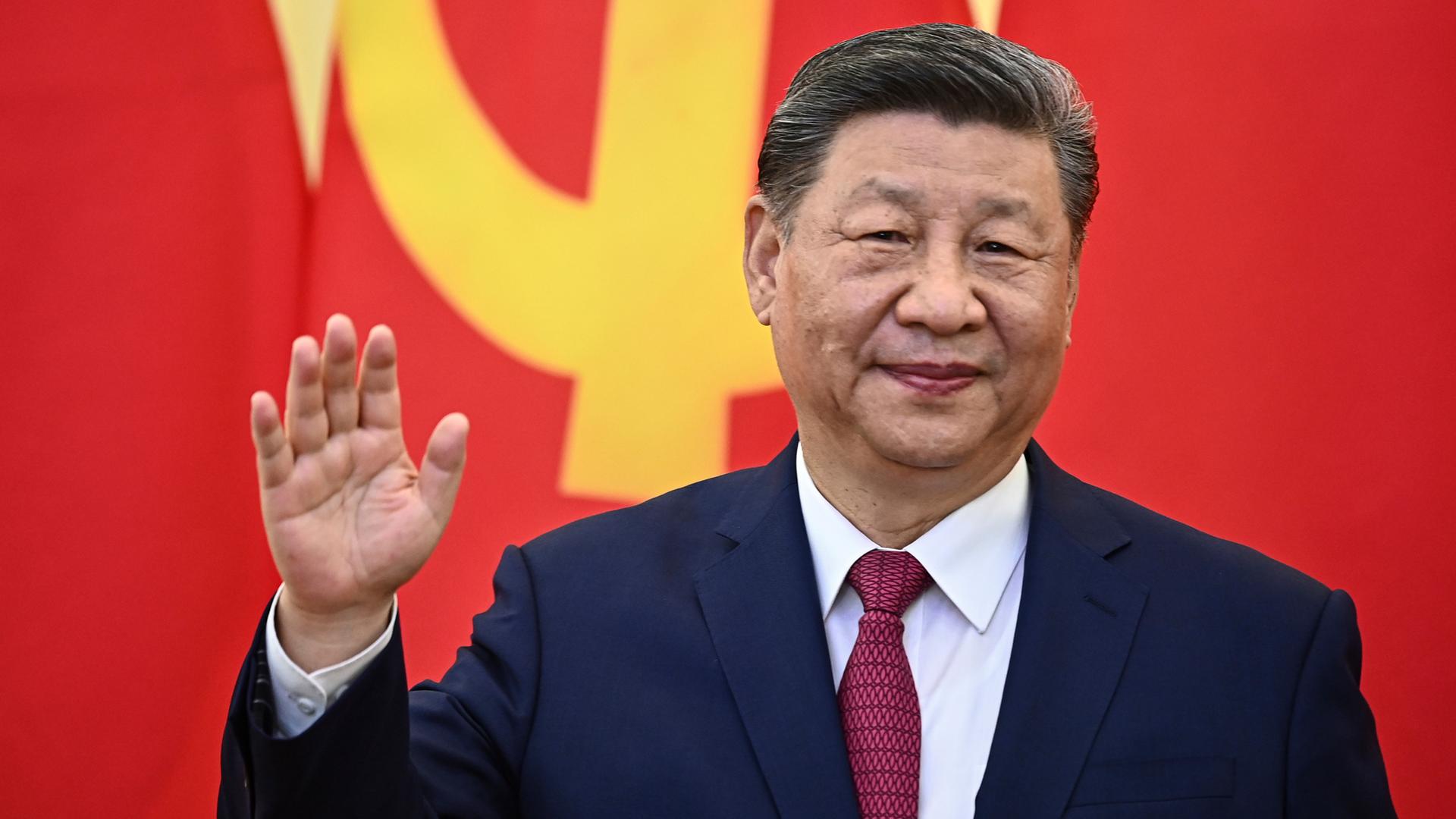 Xi Jinping steht vor einer roten Flagge mit gelbem Hammer und Sichel-Aufdruck und hebt leicht die Hand.