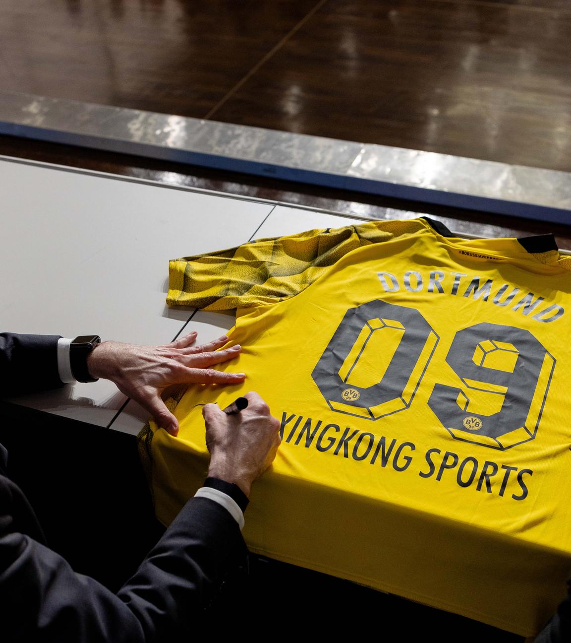 Zwei Männer bei Vertragsunterzeichnung von XingKong Sports mit Borussia Dortmund: Sie unterschreiben mit Filzstiften auf einem Dortmund-Trikot