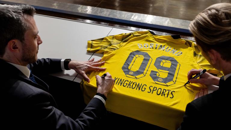 Zwei Männer bei Vertragsunterzeichnung von XingKong Sports mit Borussia Dortmund: Sie unterschreiben mit Filzstiften auf einem Dortmund-Trikot
